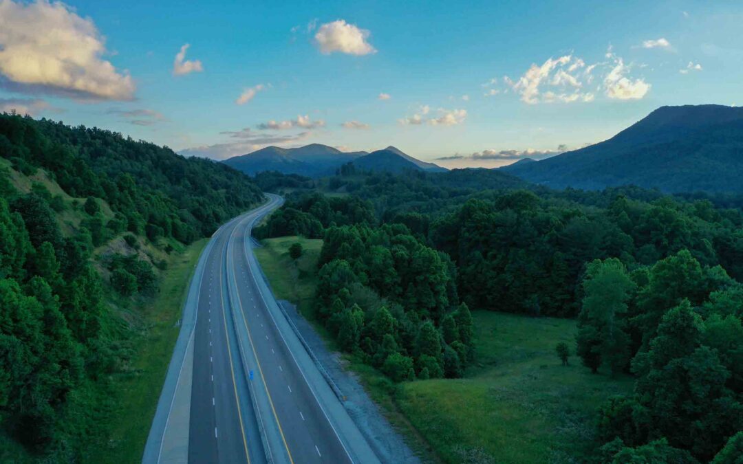 A winding road cuts through lush, green forests under a clear blue sky with fluffy clouds - tps://www.widenerins.com